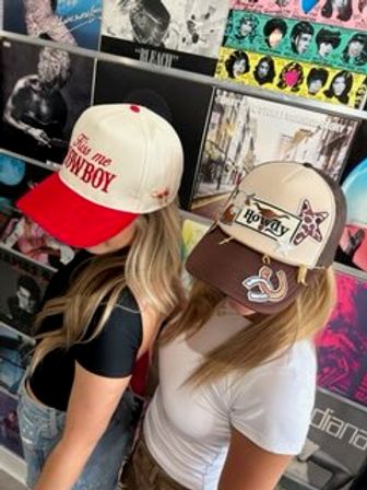 Two women wearing trendy trucker caps — a white-and-red cap with bold red lettering and a beige-brown cap with a “Howdy” patch and star — standing in front of a colorful wall of vinyl album covers in a record store