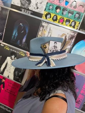 Wide-brimmed powder-blue hat with navy-and-cream ribbon, pearl trim, feathers and a tucked vintage photo, worn against a colorful vintage vinyl record wall in a retro record shop.