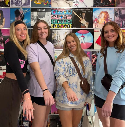 Four smiling friends pose in front of a colorful wall of vinyl record covers, extending their hands to show matching bracelets while wearing casual outfits and crossbody bags.