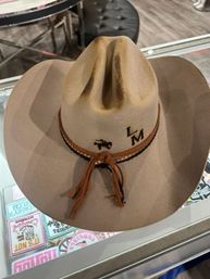 Tan felt western cowboy hat with pinched crown, braided leather band and tassels, black horse silhouette and "LM" lettering on the crown, displayed on a glass counter in a shop.