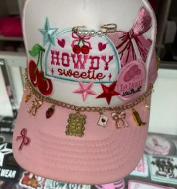 Pink trucker cap with embroidered "HOWDY sweetie" patch, cherries, stars and bow accents, decorated with a gold charm chain of tiny trinket charms