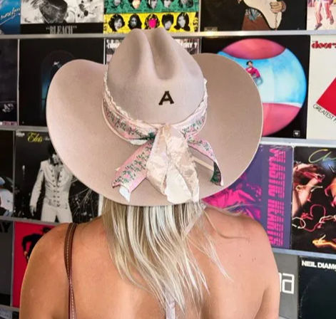 Back view of a person with long blonde hair wearing a beige cowboy hat monogrammed “A” tied with a floral scarf, standing before a colorful wall of vinyl album covers.