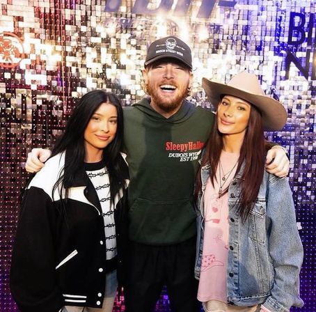 Smiling trio posing in front of a sparkly sequined event backdrop: a man in a cap and green hoodie with arms around two women—one in a black-and-white jacket, the other in a wide-brimmed hat and denim jacket.