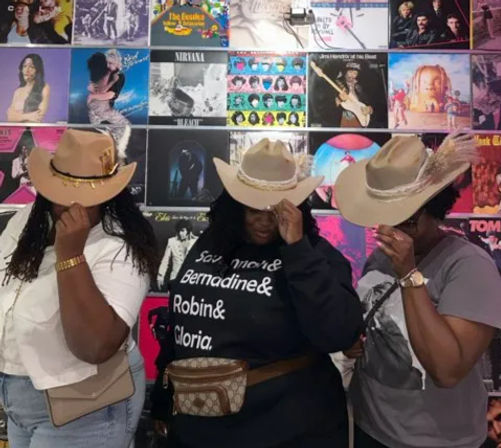 Three people wearing beige cowboy hats tip their brims while posing in front of a colorful wall of vinyl album covers in a record-store display.