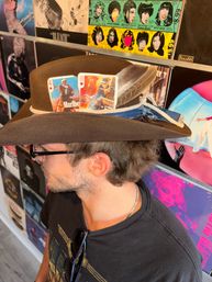 Person wearing a brown felt cowboy hat topped with two illustrated playing cards, a feather and a denim ribbon, posed in front of a colorful wall of vinyl album covers.