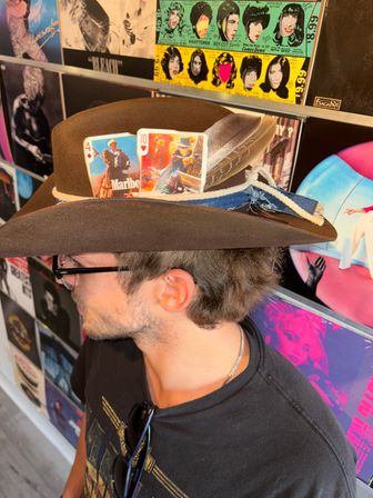 Person wearing a brown felt cowboy hat topped with two illustrated playing cards, a feather and a denim ribbon, posed in front of a colorful wall of vinyl album covers.