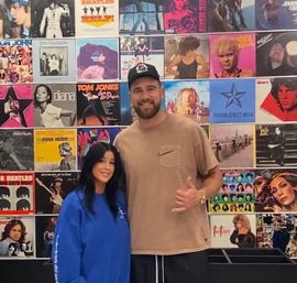 Two people smiling and posing in front of a colorful wall of vinyl album covers inside a music-themed space; man in a tan shirt and cap makes a shaka gesture while a woman in a blue sweatshirt stands beside him.