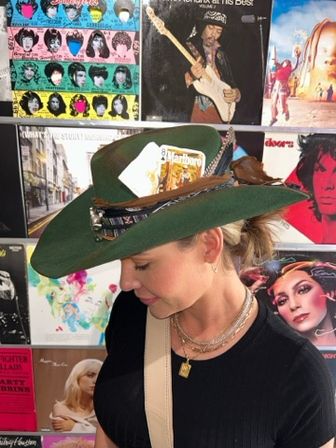 Side-profile of a woman wearing a green felt cowboy hat adorned with feathers, a playing card and cigarette pack in the hatband, layered necklaces and a black top, posed against a wall of vintage vinyl album covers in a record-store style display.