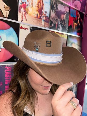 Person playfully tipping a brown felt cowboy hat with a white-and-blue band and turquoise charm, hand showing stacked rings, in front of a colorful wall of vinyl album covers.