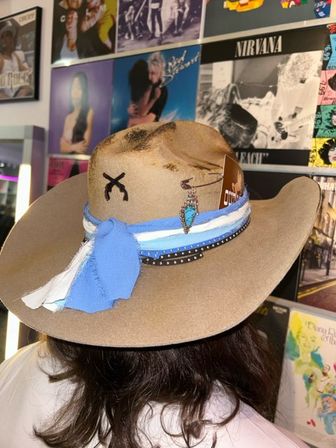 Rear view of a person wearing a tan cowboy hat with a blue-and-white fabric band, turquoise pendant and studded trim, photographed against a wall of colorful music posters and vinyl records in a record shop.