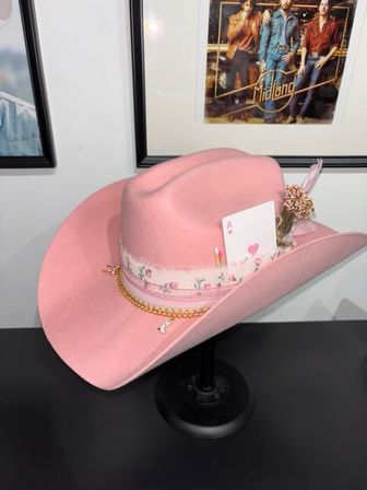 Pink felt cowboy hat on a black stand with a floral ribbon and gold chain trim, decorated with an Ace of Hearts card and dried flowers — western accessory tabletop display.
