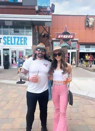 Couple wearing cowboy hats and sunglasses holding plastic drinks, posing outside an outdoor concert venue entrance with large signage; woman in white crop top and pink flared jeans, man in white t‑shirt and black pants.