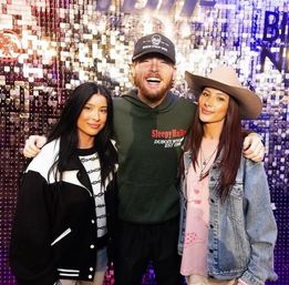 Three smiling adults posing for a group photo against a sparkly sequin event wall: bearded man in a baseball cap and green hoodie stands center with arms around two women — one in a black-and-white jacket and the other in a denim jacket and tan wide-brim hat.