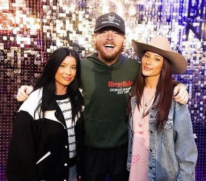 Three smiling adults posing for a group photo against a sparkly sequin event wall: bearded man in a baseball cap and green hoodie stands center with arms around two women — one in a black-and-white jacket and the other in a denim jacket and tan wide-brim hat.