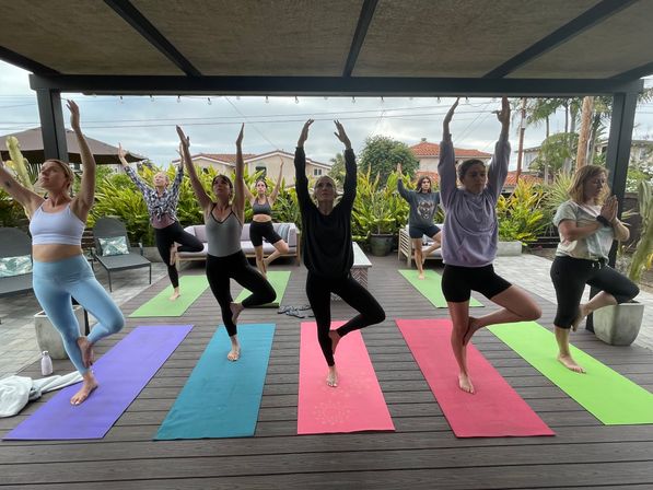 Seven people balancing in tree pose on colorful yoga mats during an outdoor group yoga class on a covered wooden deck in a residential backyard