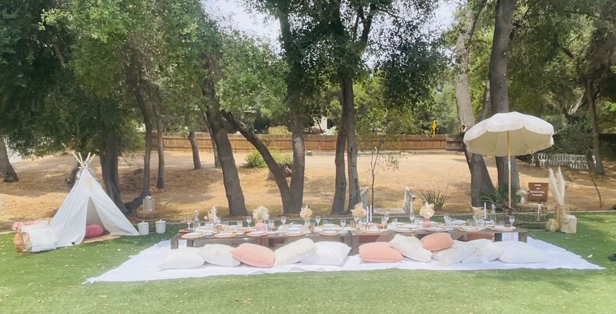Boho outdoor picnic in a shaded park-like yard with low wooden tables on a white blanket, pastel pink and white cushions, place settings and floral centerpieces, candles, a small teepee on one side and a parasol on the other beneath oak trees.
