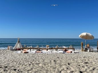 Boho seaside picnic on a sandy beach with low wooden tables, floor cushions, a small teepee and fringed umbrella facing the calm blue ocean under a clear sky with a seagull overhead.