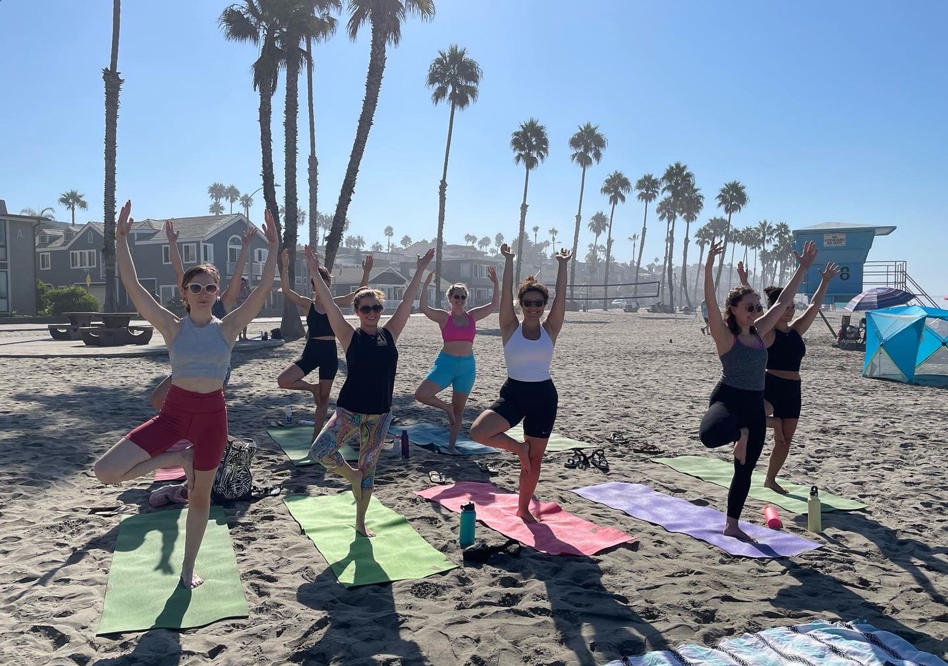 Group beach yoga class practicing tree pose on colorful mats on a sunny sandy beach with tall palm trees and a lifeguard tower in the background