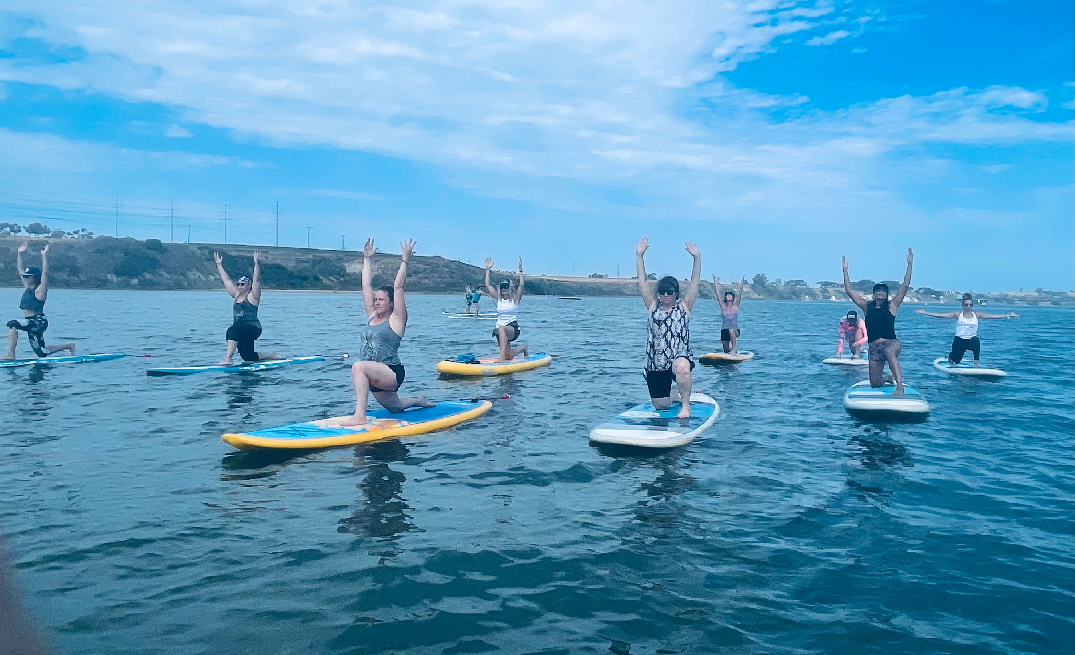 Group paddleboard yoga on a calm coastal bay, participants on colorful stand-up paddleboards kneeling with arms raised against a blue sky and distant shoreline