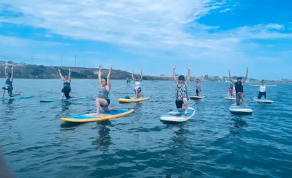 Group paddleboard yoga on a calm coastal bay, participants on colorful stand-up paddleboards kneeling with arms raised against a blue sky and distant shoreline
