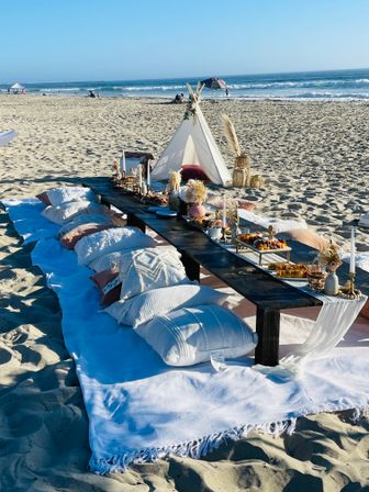 Boho-style beach picnic on sandy oceanfront — low dark-wood table surrounded by white pillows and blankets, small teepee, charcuterie boards, candles and dried floral decor with waves and clear blue sky in the background