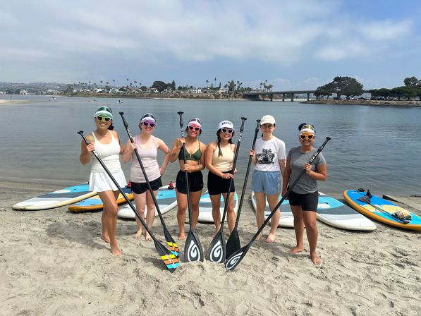 Six friends in sun hats and sunglasses holding stand-up paddleboard paddles on a sandy beach beside a calm bay, with paddleboards on the shore, palm trees and a bridge in the distance.
