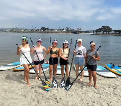 Six friends in sun hats and sunglasses holding stand-up paddleboard paddles on a sandy beach beside a calm bay, with paddleboards on the shore, palm trees and a bridge in the distance.