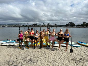 Nine women in colorful swimwear posing with stand-up paddleboard paddles on a sandy beach, paddleboards behind them and a palm-lined bay with a bridge under a cloudy sky.