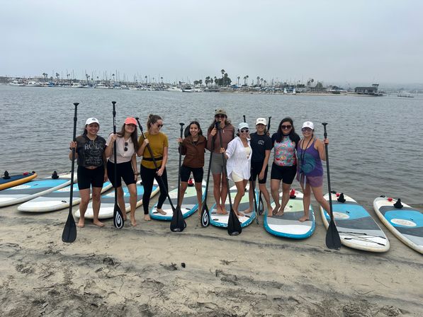 Nine people posing on paddleboards at a sandy beach near a marina, holding paddles with sailboats and palm trees in the background under an overcast sky.