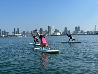 Three people doing stand-up paddleboard (SUP) yoga handstand poses on calm bay water with the San Diego skyline and sailboats under a clear blue sky