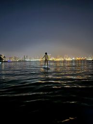 Silhouetted paddleboarder on a calm harbor at night with a glowing city skyline and golden lights reflecting on rippling water.