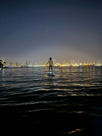 Silhouetted paddleboarder on a calm harbor at night with a glowing city skyline and golden lights reflecting on rippling water.