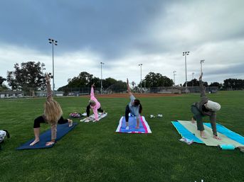 Four people doing outdoor yoga on colorful mats on a green park field beside a baseball diamond, stretching into low lunges with arm reaches under an overcast sky