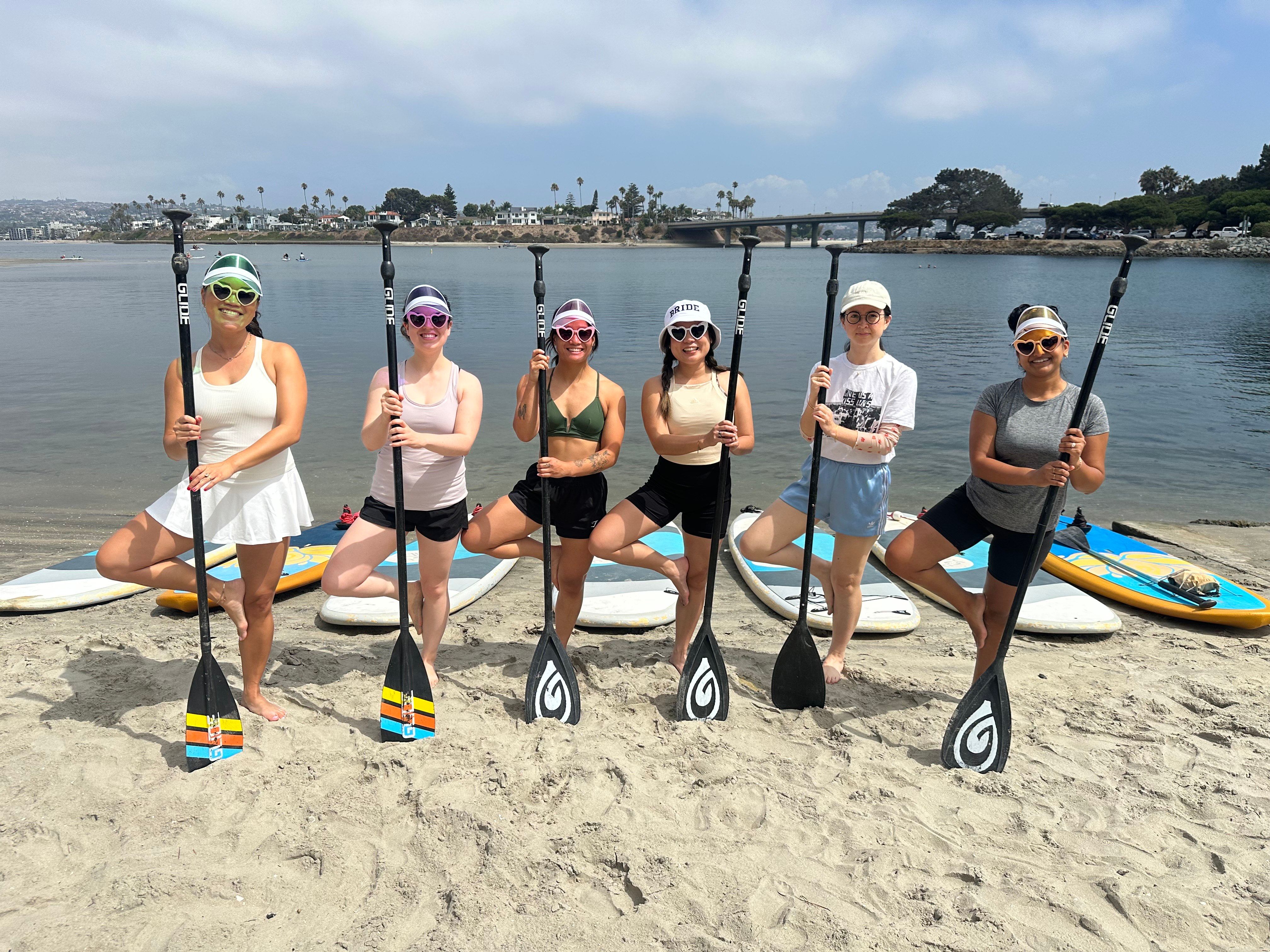 Six people in colorful sunglasses posing with stand-up paddleboards and paddles on a sandy beach, calm bay with palm trees and a bridge in the background.