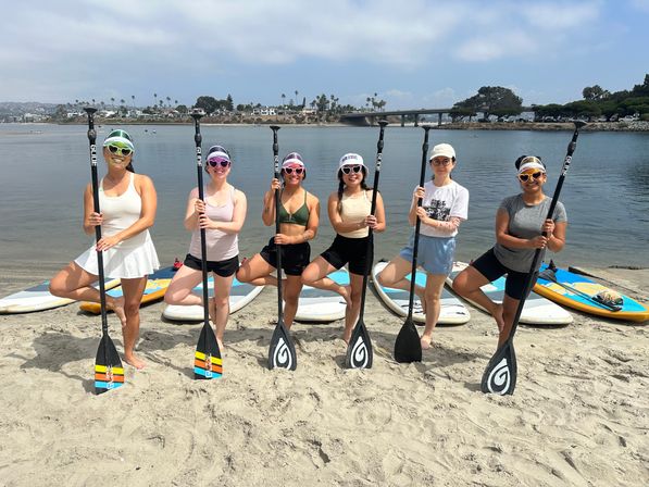 Six people in colorful sunglasses posing with stand-up paddleboards and paddles on a sandy beach, calm bay with palm trees and a bridge in the background.
