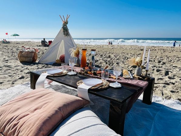 Sunlit beach picnic on a sandy shore with a low wooden table, charcuterie spread, glassware, candles, cushions and a small teepee by the ocean waves.