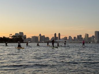 Silhouetted stand-up paddleboarders on San Diego Bay at sunset with palm trees and the downtown skyline in the background