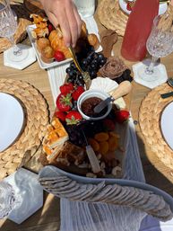 Sunlit outdoor brunch charcuterie board on a wooden patio table with strawberries, black grapes, dried apricots, mixed nuts, cheese wheel, jam, crackers, waffles and pastries, hand reaching with a gold fork.
