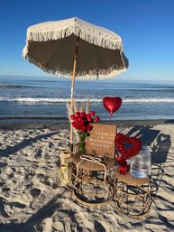 Sunny sandy beach scene with a fringed umbrella over wicker tables holding a vase of red roses, heart balloon, water dispenser and a wooden sign reading Yoga in Beautiful Places beside the ocean