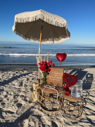 Sunny sandy beach scene with a fringed umbrella over wicker tables holding a vase of red roses, heart balloon, water dispenser and a wooden sign reading Yoga in Beautiful Places beside the ocean