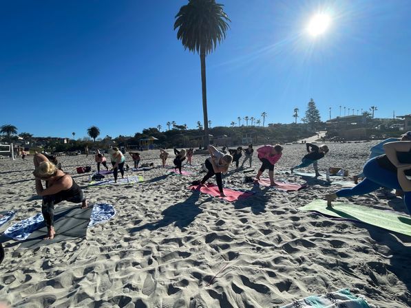 Sun-drenched beach yoga class on a sandy shore, people stretching on colorful mats beneath a tall palm tree with clear blue sky and coastal buildings in the background