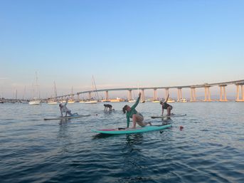Group of paddleboarders doing yoga on calm San Diego Bay with sailboats and the Coronado Bridge in the background at sunset