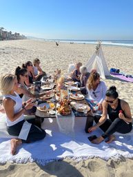 Group of friends enjoying a seaside beach brunch around a low wooden table with pastries, drinks and a small teepee on a sunny sandy shore