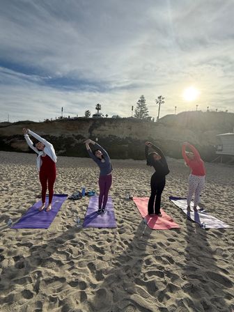 Four people doing side-stretch yoga on colorful mats on a sandy beach at sunset with coastal cliffs and palm trees in the background