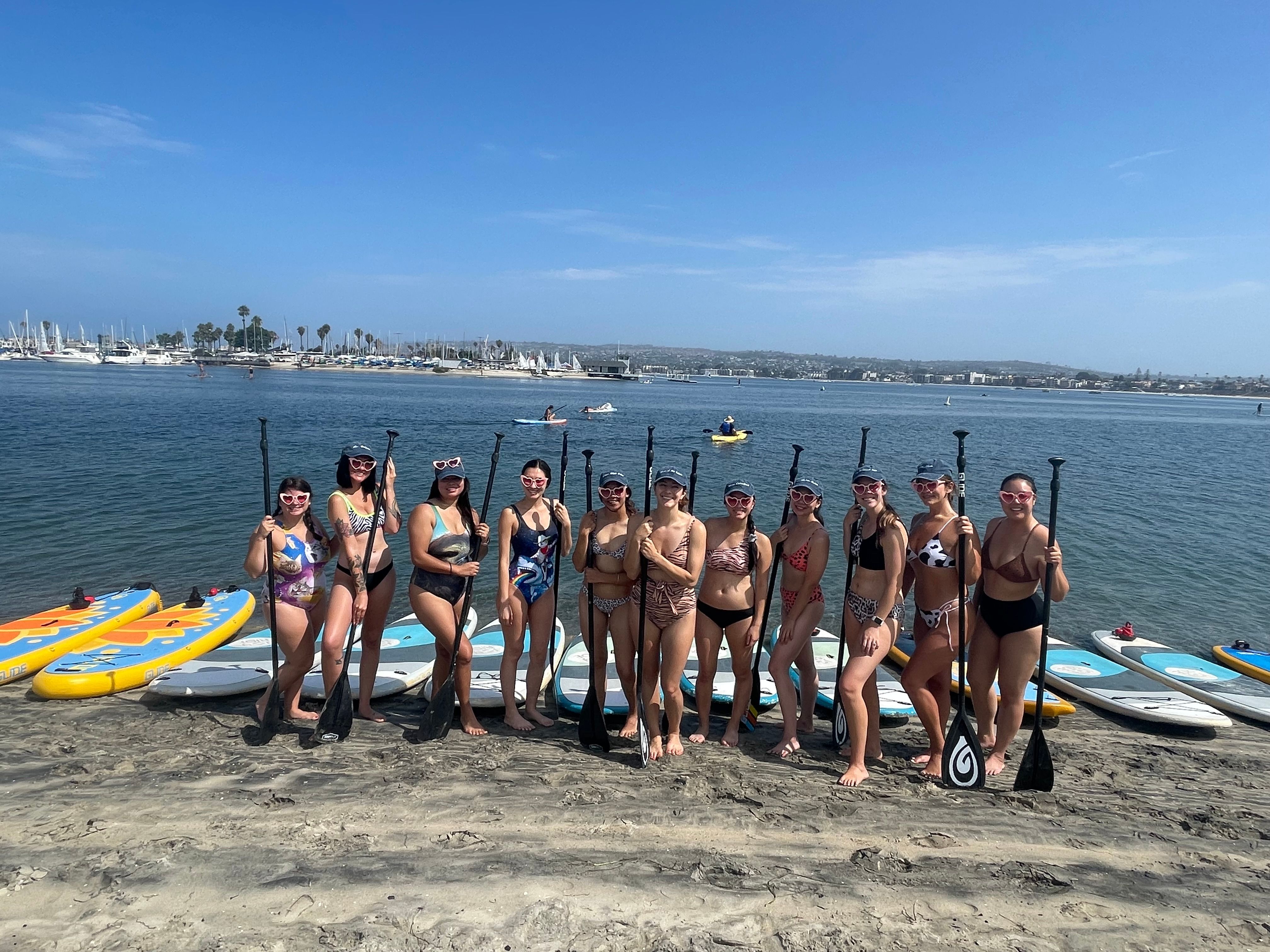 Group of paddlers in swimsuits posing on a sandy shore with paddles and colorful paddleboards, calm bay and marina with sailboats under a bright blue sky