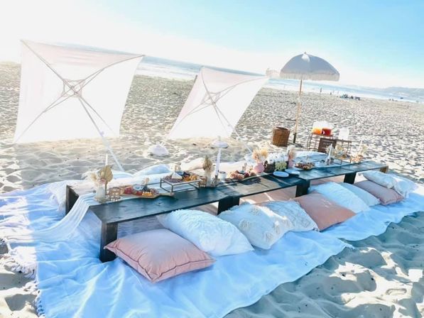 Sunlit seaside picnic on a sandy beach with a low dark wooden table, pastel pillows and white canopy umbrellas, charcuterie and decorative vases with the ocean in the background