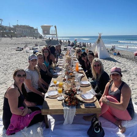 Long low wooden table set for an oceanfront beach brunch with a large group of women seated on blankets, fruit platters and mimosas on the sand, sunny blue sky, lifeguard tower and small teepee by the shore.