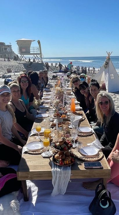 Long low wooden table set for an oceanfront beach brunch with a large group of women seated on blankets, fruit platters and mimosas on the sand, sunny blue sky, lifeguard tower and small teepee by the shore.