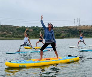 Group of four people practicing stand-up paddleboard yoga on colorful inflatable boards in a calm coastal estuary with rolling grassy hills and an overcast sky.