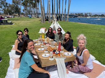 Women enjoying a sunny boho-style picnic on blankets and cushions at a waterfront park with palm trees and a bridge, gathered around a low wooden table set with plates, pastries, fruit, charcuterie and pitchers.